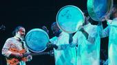4 men on stage, one plays a guitar and three are holding percussion instruments in the air.