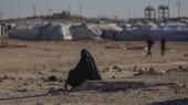 A woman sits in the foreground. In the background tents of a camp are visible.