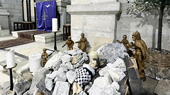 Eine Puppe liegt eingewickelt in ein weiß-schwarz kariertes Tuch, umgeben von Kerzen und Trümmern in einer Kirche