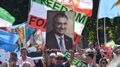 A man in a crowd holds up an image of Crown Prince Reza Pahlavi. There are many flags in the background, including the flag of Iran.