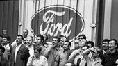 Schwarz-weiß Bild von Männern, die vor einer Wand stehen. Auf der Wand ist das Firmenlogo von Ford zu sehen.