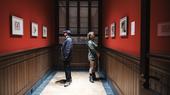 Two people in a corridor, facing away from eachother and looking at paintings hanging on the walls.
