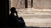 A silhouetted woman sits in the doorway of a ruined building looking out onto the street.