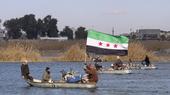 Ein Boot fährt über einen Fluss. Eine Passagierin hält die syrische Nationalflagge in den Händen. 