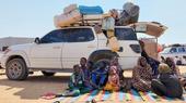 A family sit on a blanket on the ground in front of a truck loaded with bags and suitcases.
