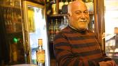 Ein Mann im gestreiften Pullover, mit weißem Schnauzbart und Haar, steht in einer Bar.