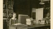 A black and white image of a young woman sitting in a room full of books.