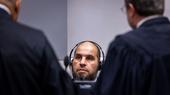 A man is sitting with headphones on; in front of him stand two men in lawyers’ robes, with their backs to the camera.
