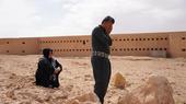 A man holds his head in his hands. Behind him a woman is sitting on the ground. In the background there is a large prison building.