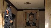 A man in uniform, holding a rifle, seen from behind. He is looking at a damaged mural of Bashar al-Assad.