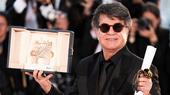 A man wearing a black suit and sunglasses holds an award and a scroll.