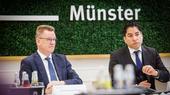 Zwei Männer in Anzug sitzen an einem Konferenztisch, im Hintergrund an der Wand der Schriftzug "Münster"