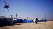 A woman and a child walk towards a cluster of tents. A Batman shaped kite is visible in the sky.