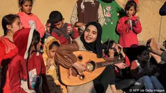 A smiling woman in a black headscarf plays the oud as she is surrounded by a group of children, some of whom are clapping along to the music