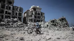 A man pushes a bicycle through rubble. In the background are three destroyed buildings.