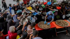 Eine große Gruppe von Menschen mit leeren Tellern, Schüsseln und anderen Gefäßen drängt sich um einen Helfer, der Essen aus einer großen Pfanne ausgibt.