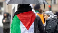 One person stands with their back to the camera, carrying an umbrella and with a Palestinian flag around their shoulders.