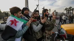 A group of young men carrying guns, one has a Syrian revolutionary flag wrapped around his shoulders.