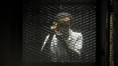 A man behind a wire panel holds his hands up to mimic taking a photograph.