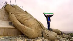 A man with the flag of free Syria stands on the head of a toppled Assad statue.