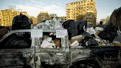 A burnt out truck full of trash bags. On the window frame a sign reads "Here lies Mubark's government!"