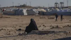 A woman sits in the foreground. In the background tents of a camp are visible.