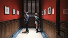 18th Istanbul Biennial (Photo: IKSV | Mete Kaan Özdilek) Two people in a corridor, facing away from eachother and looking at paintings hanging on the walls.