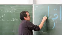 Ein Mann mit Brille schreibt auf Arabisch an eine grüne Tafel.
