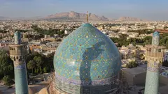 A mosque dome with green mosaic tiles against the backdrop of a city, with mountains in the background.