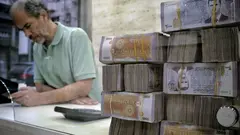 A man stands at a desk looking at his phone. In the foreground, a stack of 2000 SYP notes bearing the face of Bashar al-Assad.
