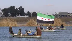 Ein Boot fährt über einen Fluss. Eine Passagierin hält die syrische Nationalflagge in den Händen. 