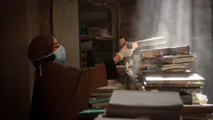 A woman wearing a hijab and a face mask dusts books in a destroyed library.