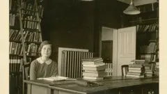 A black and white image of a young woman sitting in a room full of books.