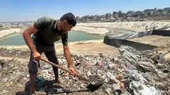 A man with a short beard is holding a shovel; within sight are a pool of water and some tents that have been pitched