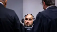 Ein Mann sitzt mit Kopfhörern, vor ihm stehen zwei Männer in Anwaltsroben mit dem Rücken zur Kamera.