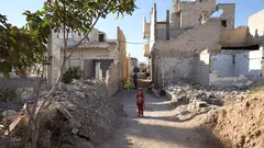 Ein Mädchen steht auf einem Weg zwischen kaputten Gebäuden und Schutt. Im Hintergrund ein weiteres Mädchen und ein alter Mann.