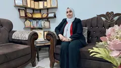 سيدة ترتدي الحجاب وتجلس على أريكة وخلفها رف من الدروع.