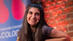 Eine Frau mit langen braunen Haaren und einem Mikrofon lehnt in einem Sessel und lächelt in die Kamera.
