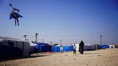 A woman and a child walk towards a cluster of tents. A Batman shaped kite is visible in the sky.