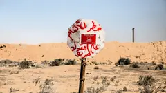 Ein Schild auf Arabisch warnt vor Minen, im Hintergrund eine trockene, flache Landschaft.