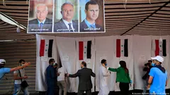 لجنة اقتراع لمواطنين سوريين في لبنان في خيمة. مقصورات الاقتراع معزولة بستارات معلقة عليها الأعلام السورية. تتدلى صور المرشحين الثلاثة من السقف.