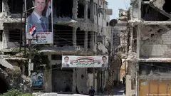 رجل يسير بين مبانٍ مدمرة في سوريا وصور رئيس النظام السوري بشار الأسد معلقة على الواجهات المكسورة.