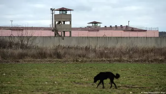 A black dog is seen walking through a field; in the background is a Turkish prison with high wall and watchtower