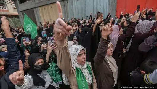 Men and women, some wearing green Hamas scarves or holding up Hamas flags, point their right index fingers in the air at a rally in April 2022