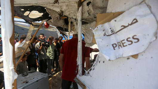 View of a destroyed tent in Gaza. In the foreground hangs a sign that reads ‘Press’ in Arabic and English. Journalists can be seen in the background.