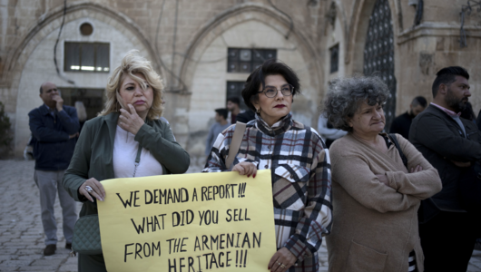 Three protesters in a square in the Old City of Jerusalem. Two of them hold a sign that reads: "We demand a report! What did you sell from the Armenian heritage?"