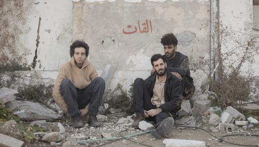 Three members of El Khat sit on the ground and on pieces of rubble in front of a white wall.