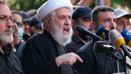 Naim Qassem wearing a white turban speaks into a microphone at a podium surrounded by men. His finger is pointing to the ground.