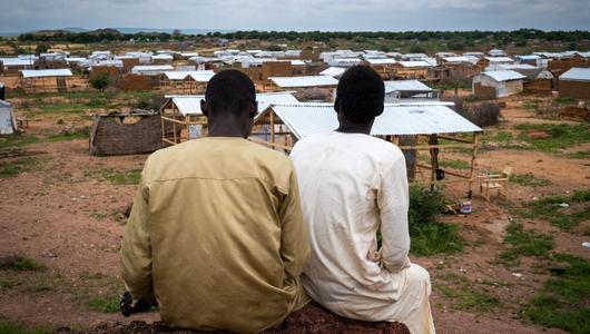 Two men sit facing away from the camera, looking over a field of makeshift shelters built from timber and corrogated iron.