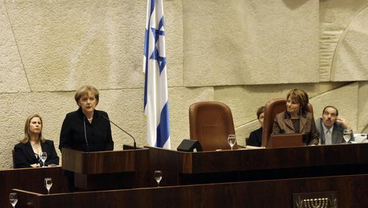 Angela merkel stands at a podium speaking into a microphone. An Israeli flag hangs in the background.